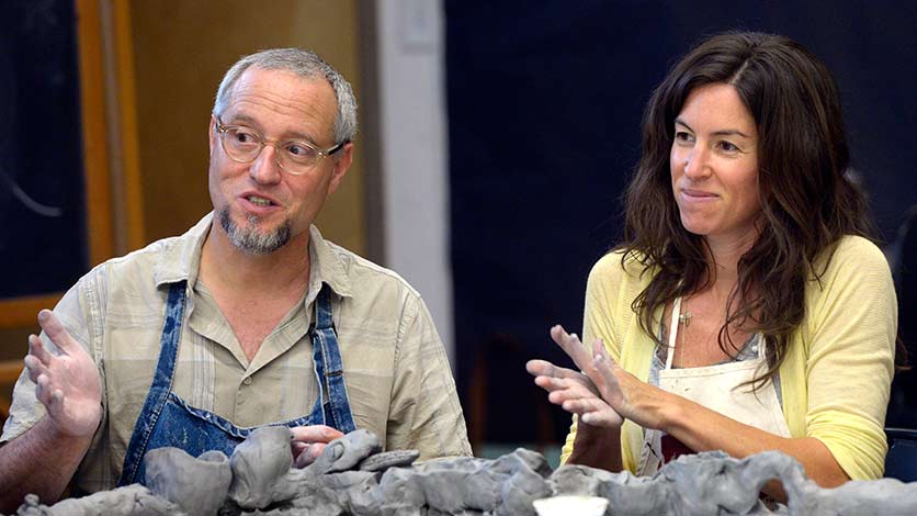 Two people sitting together side by side, only visible from the chest upwards. One is a man with short grey hair and round glasses, and the other is a woman with long, wavy dark brown hair. They are both wearing aprons and their hands are stained with clay.